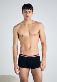 Boxeri negri, cu un brâu de marcă roșu, textură netedă, design ajustat și lungime până la mijlocul coapsei, purtați de o figură masculină.