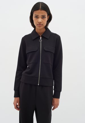 Veste noire zippée avec un col, dotée de deux poches poitrine, de poignets côtelés et d'une finition en zip argenté. Texture lisse.