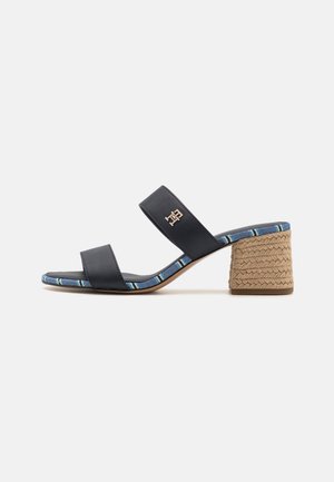 Sort sandal med to remme, flettet blokhæl og blå mønstret kant, med en lille metallisk logo på den øverste rem.