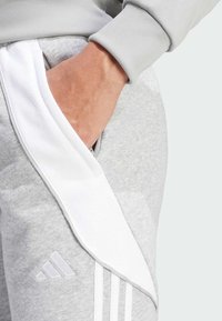 Grijze sportbroeken hebben een witte zijpaneel met drie verticale witte strepen en een Adidas-logo op de onderzijde links. Zachte textuur zichtbaar.