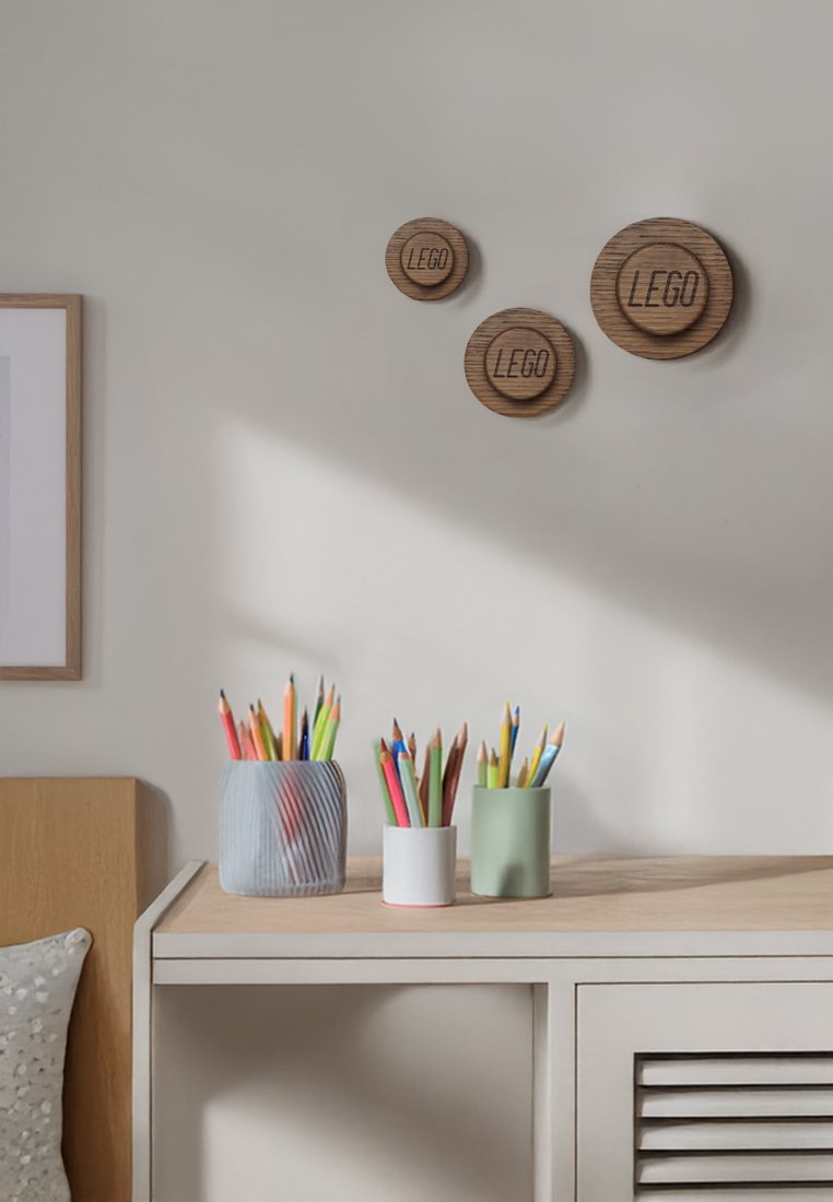 Drei Behälter mit bunten Buntstiften stehen auf einem Holztisch unter drei dekorativen hölzernen LEGO-Wandhängern.