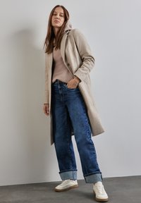 Cappotto di lana beige con colletto a punta, indossato sopra un maglione rosa chiaro. Jeans a gamba larga blu scuro con orli risvolti e sneakers bianche.