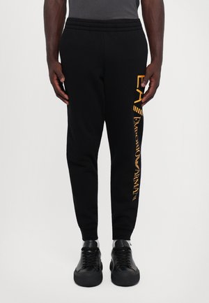 Mężczyzna noszący czarne spodnie dresowe z złotym logo Emporio Armani na prawej nodze, zestawione z czarnymi sneakersami i szarą bluzką.