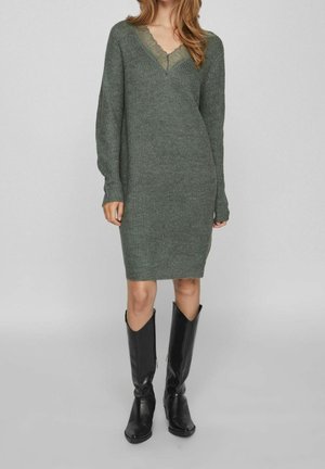 Femme portant une robe longue en maille grise avec un décolleté en dentelle, associée à des bottes en cuir noires montantes, debout sur un fond gris.