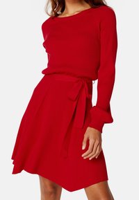 Ensemble en tricot rouge côtelé composé d'un pull et d'une jupe, avec des manches longues, un col rond et un lien à la taille sur la jupe évasée. Texture lisse.