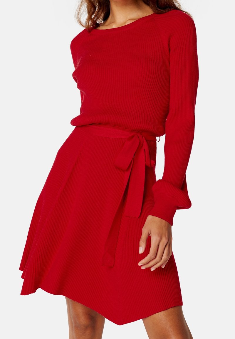 Ensemble en tricot rouge côtelé composé d'un pull et d'une jupe, avec des manches longues, un col rond et un lien à la taille sur la jupe évasée. Texture lisse.