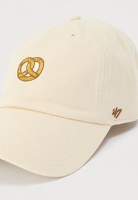 Καπέλο μπέιζμπολ κρεμ χρώματος από βαμβάκι με υφή στην επιφάνεια. Διαθέτει χρυσό κεντημένο λογότυπο πρέτζελ μπροστά και μικρό λογότυπο της μάρκας.