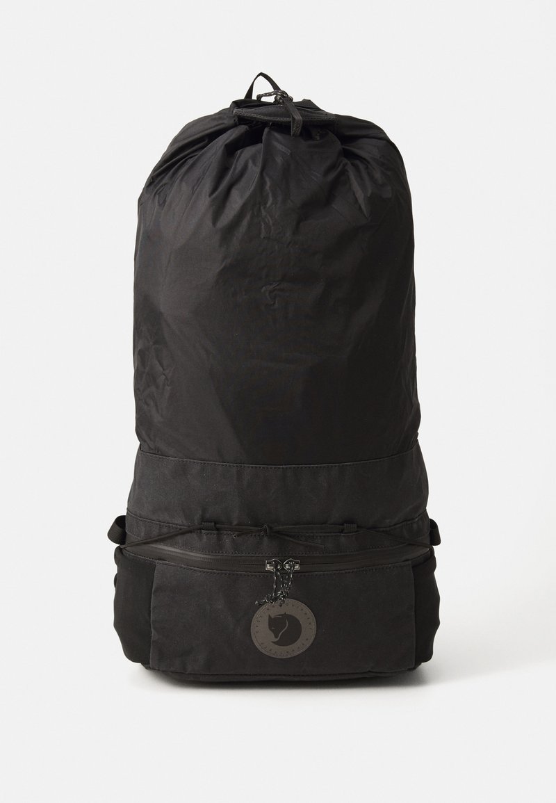 Zaino con cordino nero realizzato in tessuto resistente con una texture liscia. Presenta una tasca frontale con zip e un accentuazione decorativa a forma di logo circolare.