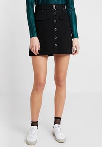 Jupe A-line noire avec des boutons sur le devant, deux poches avant et une taille ceinturée. Portée avec un haut vert et des baskets blanches. Texture lisse.