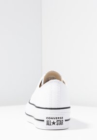 Zapatilla de lona blanca con detalles en negro, puntera redonda, diseño bajo y suela de goma. Presenta un parche de logo en el talón.