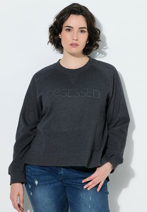 Kvinde med kort mørkt hår iført mørkegrå sweatshirt med teksten "OBSESSED" og blå revnede jeans, står op ad en lys væg.
