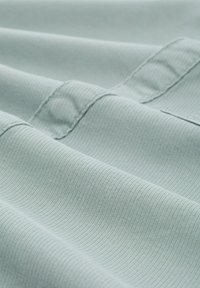 Lichtgroene ribbelstof met een zachte textuur, met zichtbare naden en stiksels, gelegd in zachte plooien.