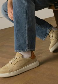 Beige suède sneakers met een witte rubberen zool en een bruin leren accent op de hiel, gecombineerd met lichtblauwe denim jeans.