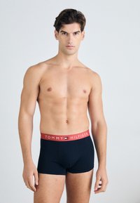 Schwarze Baumwoll-Boxershorts mit rotem Bund, der das "TOMMY HILFIGER"-Logo trägt. Das Design ist figurbetont und modern.
