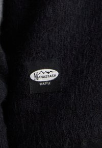 Svart stickad tyg med mjuk textur har en rektangulär etikett. Etiketten visar "Manastash" och "Seattle" mot en vit bakgrund.