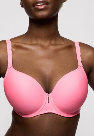 Vrouw die een roze gewatteerde beha draagt met gestructureerde bandjes en een klein hangertje in het midden aan de voorkant, tegen een effen achtergrond.