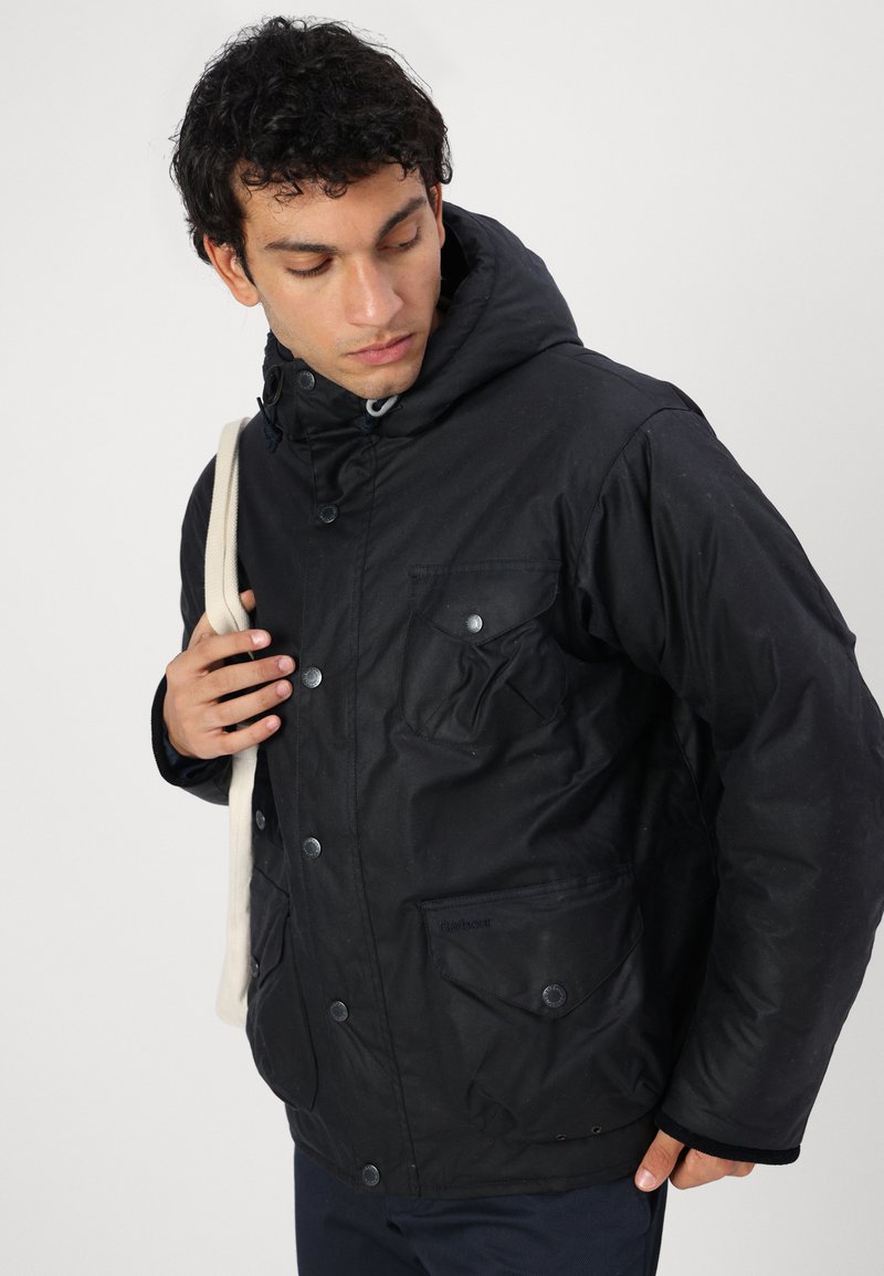Schwarze isolierte Jacke mit Kapuze, ausgestattet mit Druckknöpfen, mehreren Taschen und einer strukturierten Oberfläche. Das Model trägt einen schmalen Schulterriemen.