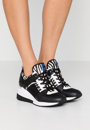 Schwarz-weiße Sneaker mit Zebramuster-Akzenten, getragen an den Füßen vor einem einfarbigen hellen Hintergrund.