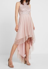 Jasnoróżowa sukienka z koralikowym, bezrękawnikowym gorsetem oraz warstwową spódnicą o kroju wysokim z przodu, niskim z tyłu. Materiał to miękki tiul o przezroczystej, lekko opadającej fakturze. Buty są w kolorze brązowym.