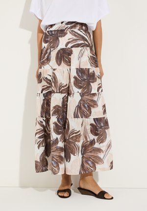 Femme portant une jupe midi à plusieurs volants avec un grand imprimé floral marron, un haut blanc et des sandales compensées noires à bout ouvert sur un fond uni.