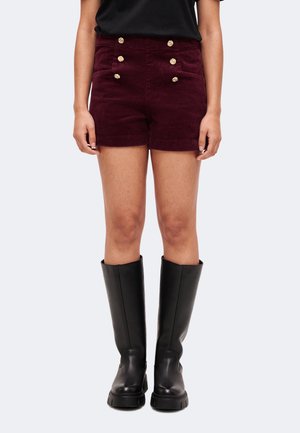 Femme portant un short bordeaux foncé avec six boutons dorés et des bottes noires montantes, debout devant un fond clair.