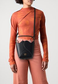 Czarna skórzana torebka typu bucket o falistym wzorze, z białym przeszyciem, złotym logo i regulowanym paskiem. Nosi ją w połączeniu z pomarańczową bluzką z długim rękawem i różowymi spodniami.