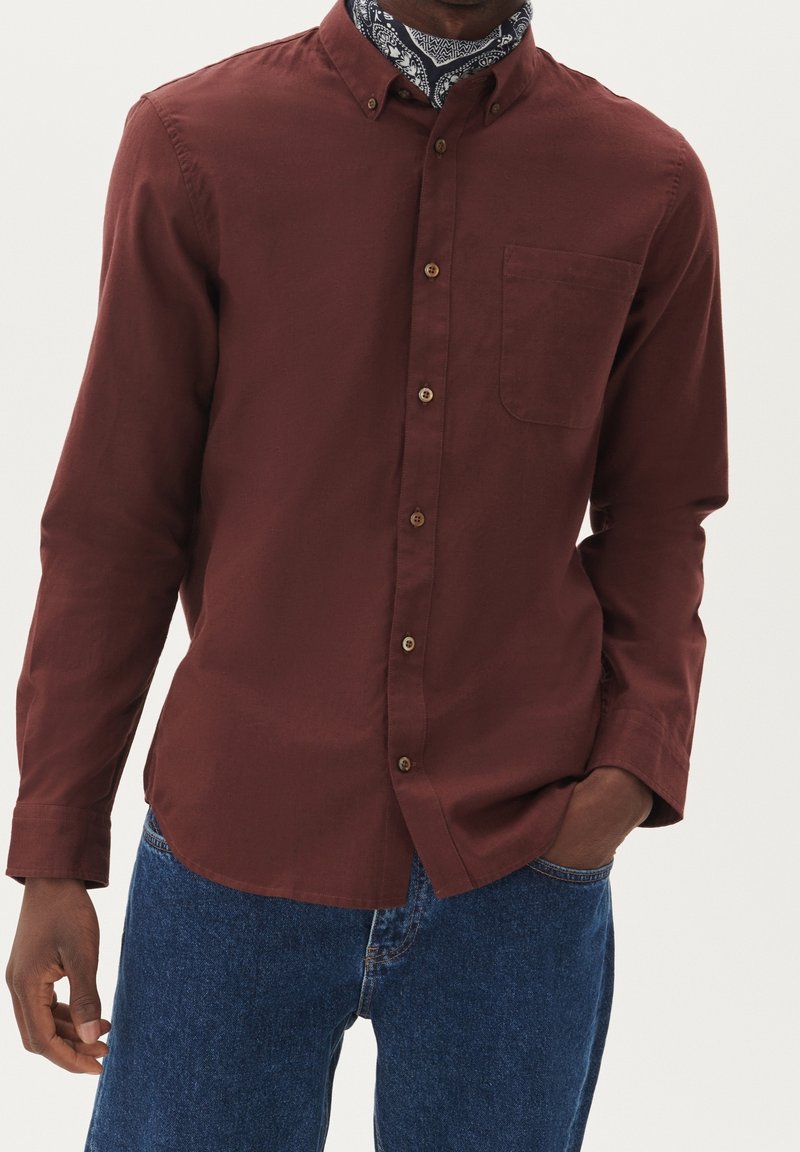 Homme portant une chemise bordeaux à boutons, un jean bleu et un foulard à motifs autour du cou, une main dans la poche, sur un fond uni.