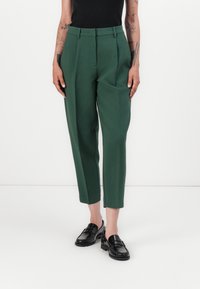 Green tailored trousers with a high waist, pleats, tapered legs, and a textured fabric, paired with black loafers. Minimalist design.