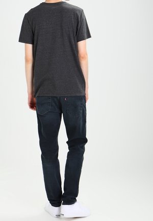 Person stående med ryggen til, iført en mørkegrå t-shirt, mørkeblå jeans og hvide sneakers mod en ensfarvet lys baggrund.