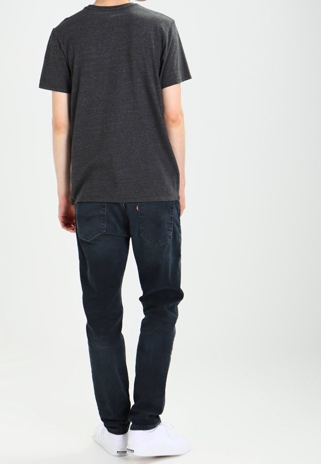 Personne debout de dos, portant un t-shirt gris foncé, un jean bleu foncé et des baskets blanches sur un fond uni clair.