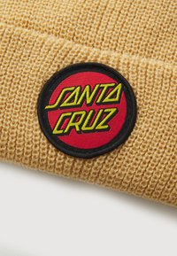 Bonnet en tricot jaune avec un patch noir rond arborant un fond rouge et "SANTA CRUZ" en texte vert. Tissu texturé avec des coutures visibles.