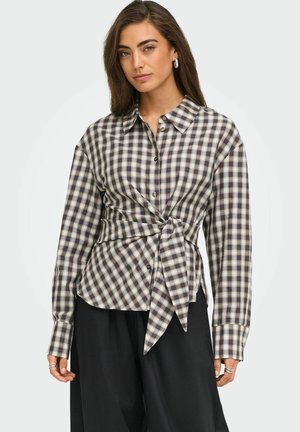 Une femme porte, sur un fond uni, une chemise à carreaux gris et crème nouée à la taille, un pantalon noir et des boucles d’oreilles argentées.