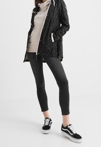 Zwarte regenjas met patroon en capuchon, beige hoodie eronder, slim-fit donkergrijze jeans en zwart-witte sneakers met witte zolen.