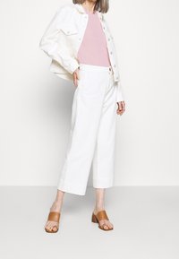 Kvinna klädd i vita cropped byxor, en ljusrosa topp, en vit knäppt jacka och bruna öppna klacksandaler som står mot en enfärgad bakgrund.