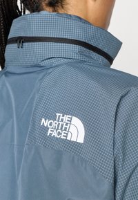 Ανοιχτό μπλε μπουφάν με σχέδιο σε πλέγμα, ψηλό γιακά και μαύρο φερμουάρ. Διαθέτει λευκό λογότυπο βουνού στην πάνω πλάτη.