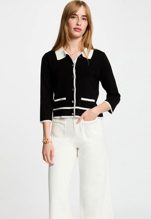 Zwarte cardigan met witte accenten, ronde halslijn, knoopsluiting, driekwartmouwen en twee voorzakken. G gedragen met witte broek.