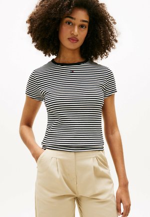 Mujer con el pelo rizado que lleva una camiseta de manga corta a rayas blancas y negras y pantalones beige, de pie con las manos en los bolsillos.