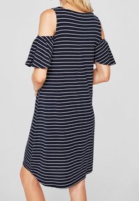 Kvinna som bär en marinblå och vit horisontell randig klänning med off-shoulder och volangärmar, står med händerna på höfterna.