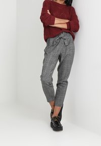 Bordowy dzianinowy sweter z teksturowanym wzorem; szare spodnie w kratę z wiązaniem w talii; czarne buty sznurowane z lekką platformą.