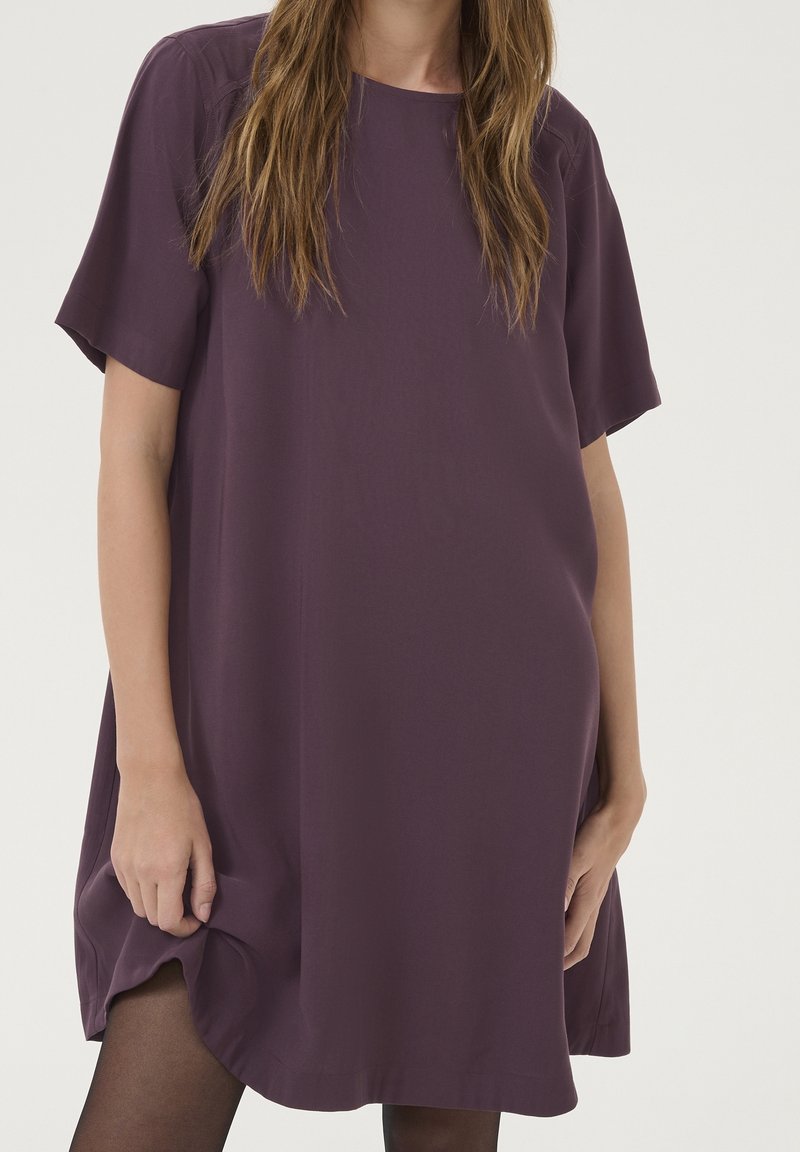 Femme portant une robe ample à manches courtes de couleur prune, debout devant un fond clair uni.