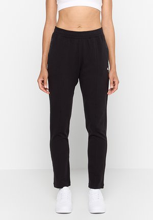 Mujer con pantalones de chándal negros ajustados, zapatillas blancas y una camiseta corta blanca, de pie frente a un fondo claro y liso.
