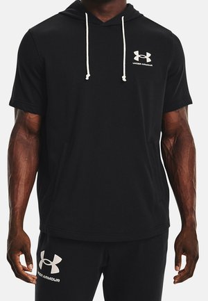 Atletyczny mężczyzna w czarnej krótkiej bluzie z kapturem Under Armour i pasujących czarnych szortach z białym logo i sznurkami.