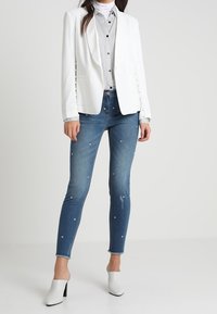Witte op maat gemaakte blazer, gestreept shirt, versierde blauwe skinny jeans met strass-steentjes, en witte hakken. Effen achtergrond.