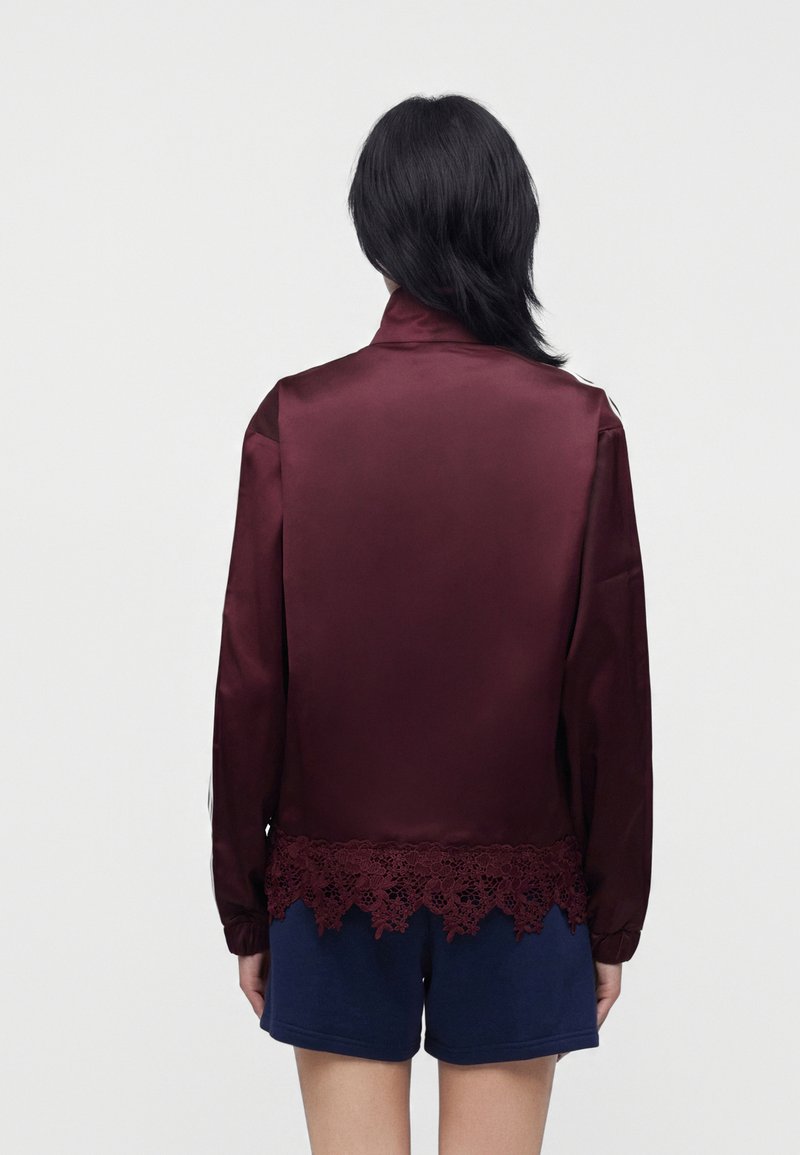 Personne aux cheveux noirs portant un haut en satin bordeaux à manches longues avec bordure en dentelle et un short bleu marine, vue de dos sur un fond uni.