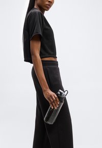 Γυναίκα με μαύρο cropped T-shirt και παντελόνι που κρατάει ένα διαφανές μπουκάλι νερού με καπάκι flip, στέκεται μπροστά σε απλό φωτεινό φόντο.