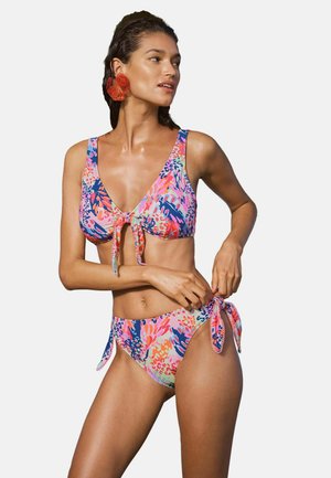 Kobieta ubrana w kolorowy bikini w kwiatowy wzór z wiązaniami, poprawia boczne wiązanie dolnej części, stoi na jednolitym tle.