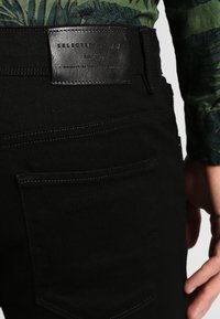 Black denim pants with a smooth texture, featuring a leather waistband label and a back pocket with contrast stitching detail.