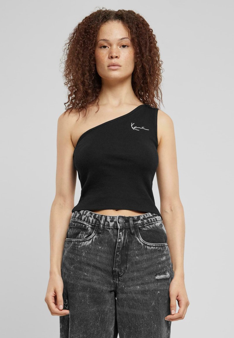 Svart ribbet crop top med én skulderdesign, med en kontrastfarge hvit logoaksent på brystet. Parret med slitte svarte jeans.