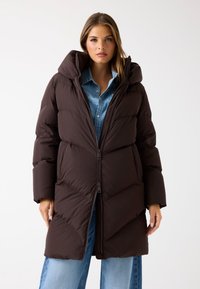 Bruine pufferjas met een capuchon, voorzien van een ritssluiting aan de voorkant, chevron-quilting en lange mouwen. G gedragen over een spijkerhemd en jeans.