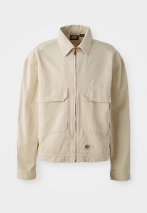 Beige, lett jakke med glidelås foran, to brystlommer og et lite logo ved nedre kant. Laget av slitesterkt bomullsstoff.